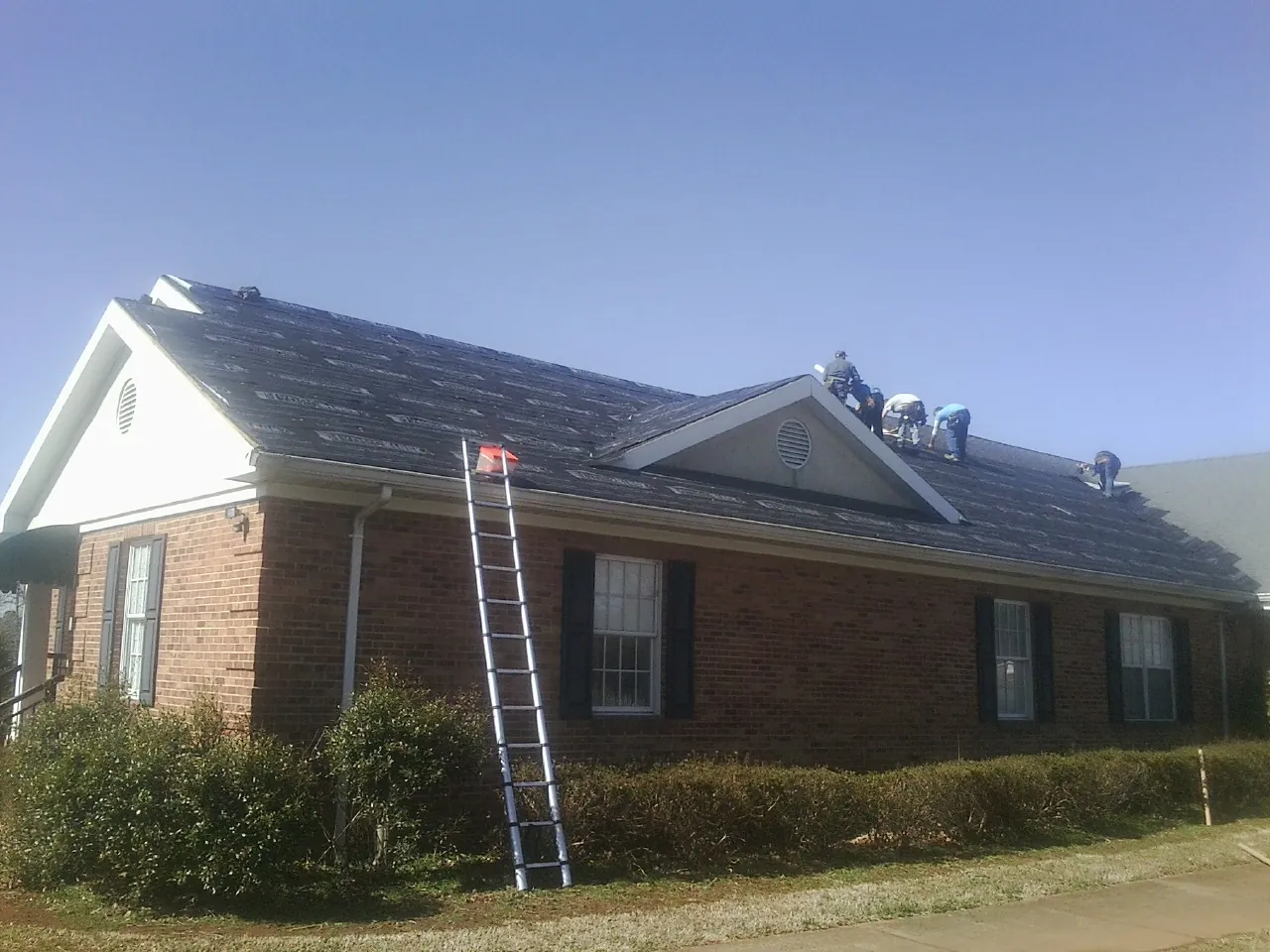 Crew tearing shingles off a brick ranch roof with a ladder set — Emergency Roof Repair work by our crew in Goddard