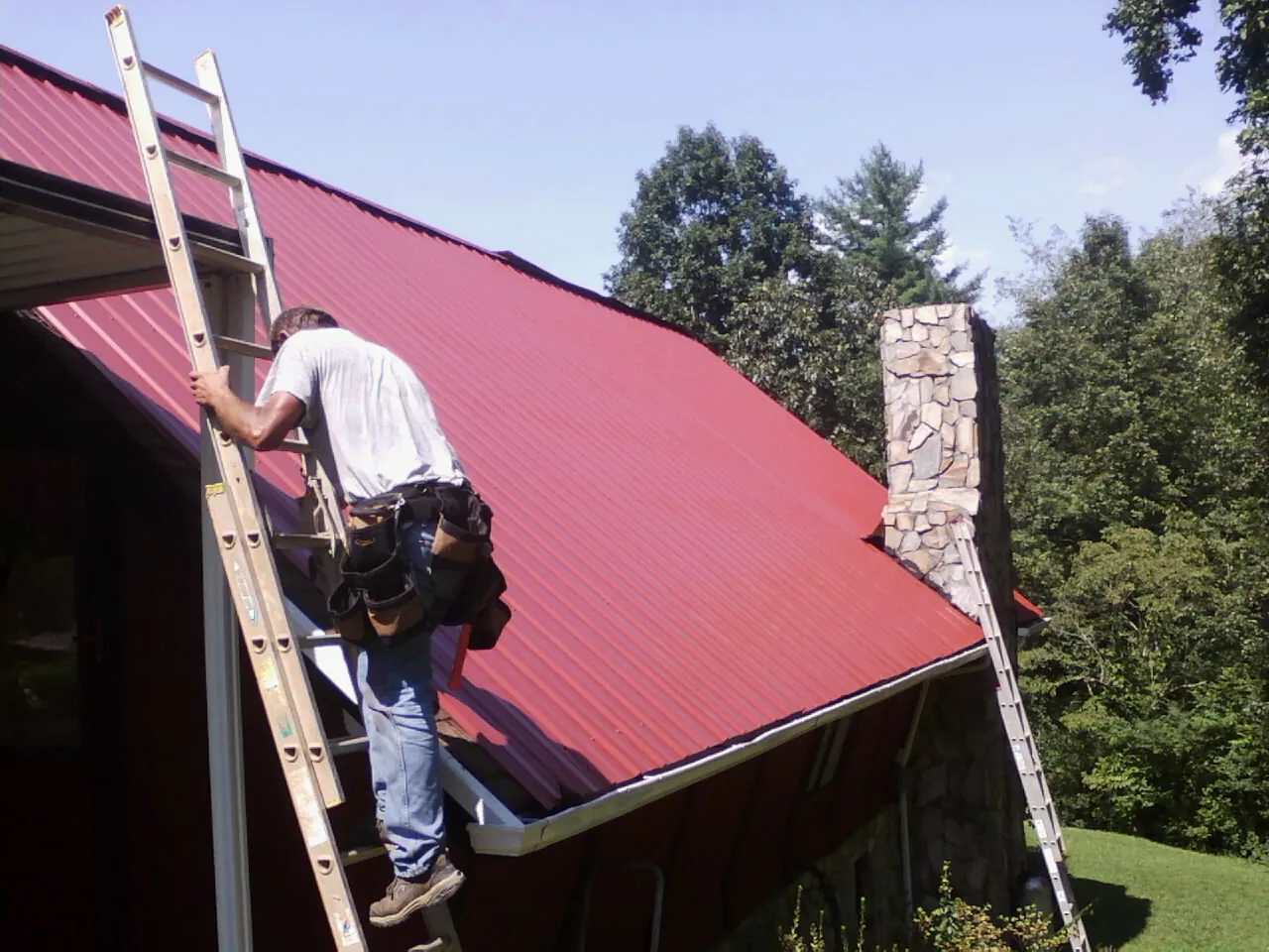 Roofer on a ladder beside a red metal roof and stone chimney — Soffit & Fascia Repair by Roofing Brothers in Goddard